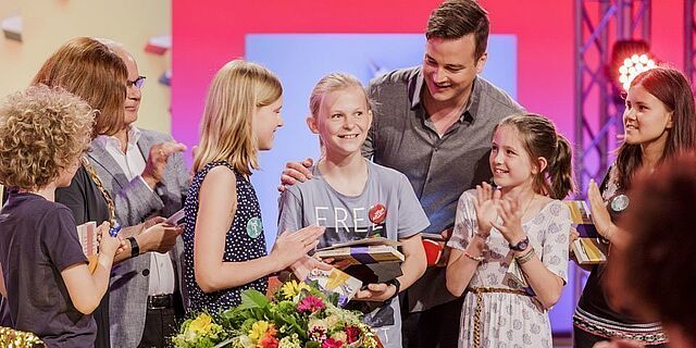 Mehrere klatschende Kinder, in der Mitte ein blonder Junge mit Pferdeschwanz, der Büchergeschenke in der Hand hält und lächelt. Ein dunkelhaariger Mann steht hinter ihm mit der Hand auf seiner Schulter und beugt sich lächelnd zu ihm nach vorn.