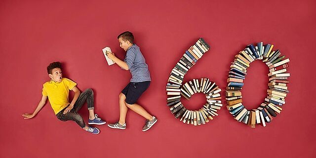 Zwei Kinder liegen auf rotem Untergrund, einer liest. Rechts neben Ihnen sind Bücher in Form der Zahl 60 angeordnet